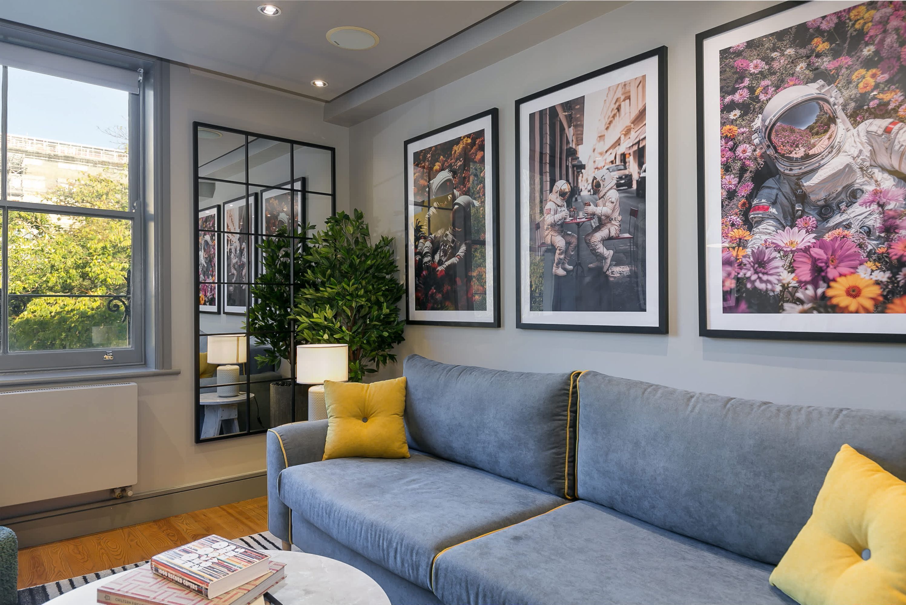 Un salon avec un canapé gris, des coussins jaunes, une table basse avec des livres, un miroir et des tableaux encadrés au mur.