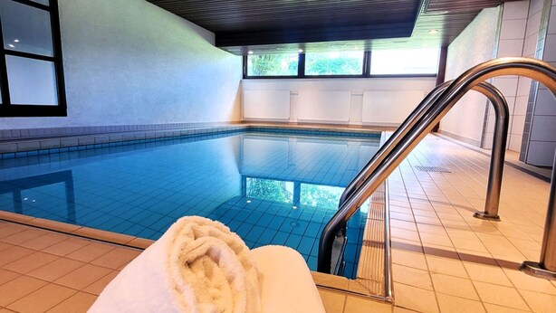 An indoor swimming pool with clear blue water, a white towel on the edge, and a view of the outside through large windows. - Apartment in Winterberg Near Ski Lift (Winterberg)