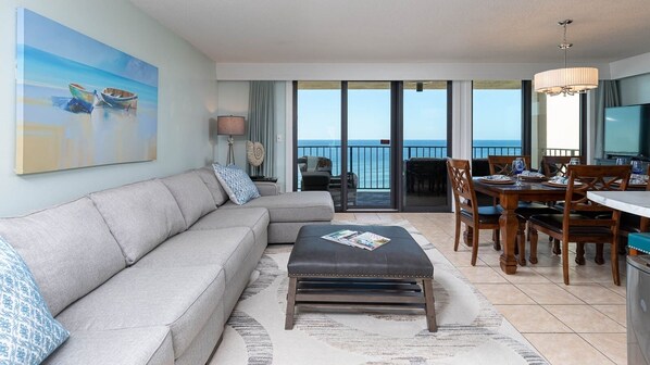 A living room with a sofa, ottoman, dining table, chairs, and a painting of boats. - Phoenix I 1071 3 Bedroom Condo (Orange Beach)
