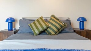 Ein ordentlich bezogenes Bett mit einer blau-weißen Tagesdecke, zwei Kissen mit grünen Streifen und zwei blaue Nachttischlampen auf Holznachttischen.
