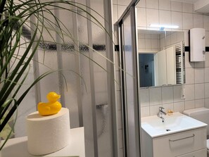 Een moderne badkamer met een douchecabine van glas, een wit wastafelmeubel en een gele rubberen eend op een rol toiletpapier.