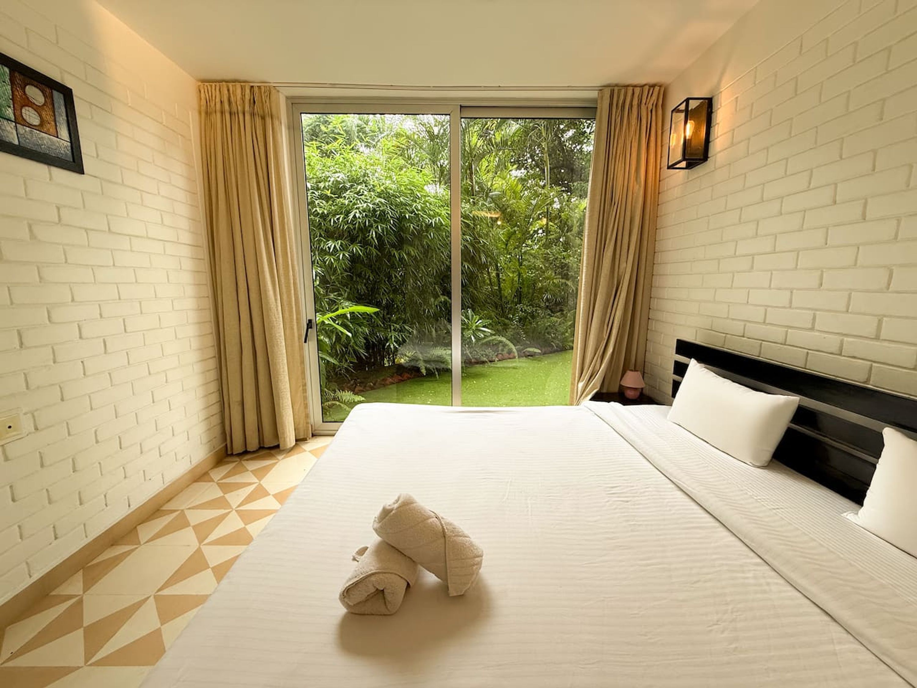 Quarto de hotel com uma cama grande, vista de um jardim exuberante através de uma porta de vidro deslizante e uma parede de tijolos brancos.