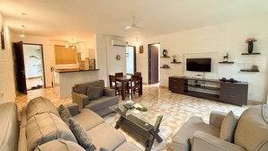 A modern living room with a sofa set, a coffee table, and a TV. There is a dining area with a table and chairs. The room has a white brick wall and a tiled floor. - Modern 2bhk w Pool Access 5km to Coco Beach (Nerul)