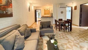 A modern living room with a sofa, a coffee table, and a dining area with chairs and a table. - Modern 2bhk w Pool Access 5km to Coco Beach (Nerul)