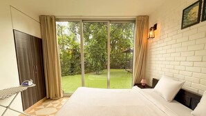 A hotel room with a large bed, a desk with an ironing board, and a view of a green garden through a sliding door. - Modern 2bhk w Pool Access 5km to Coco Beach (Nerul)