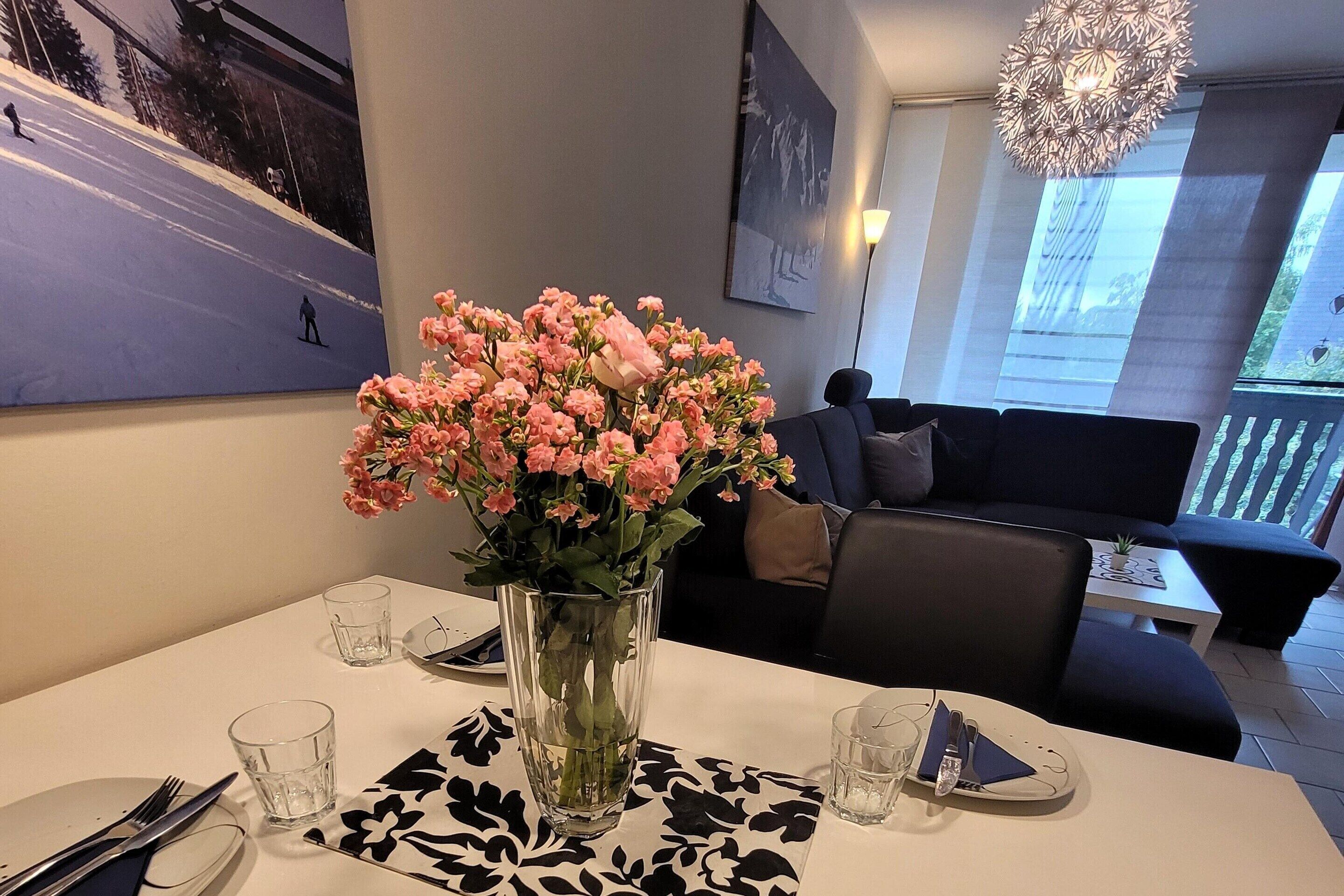 A dining area with a table set for two, a vase of flowers, and a painting on the wall.