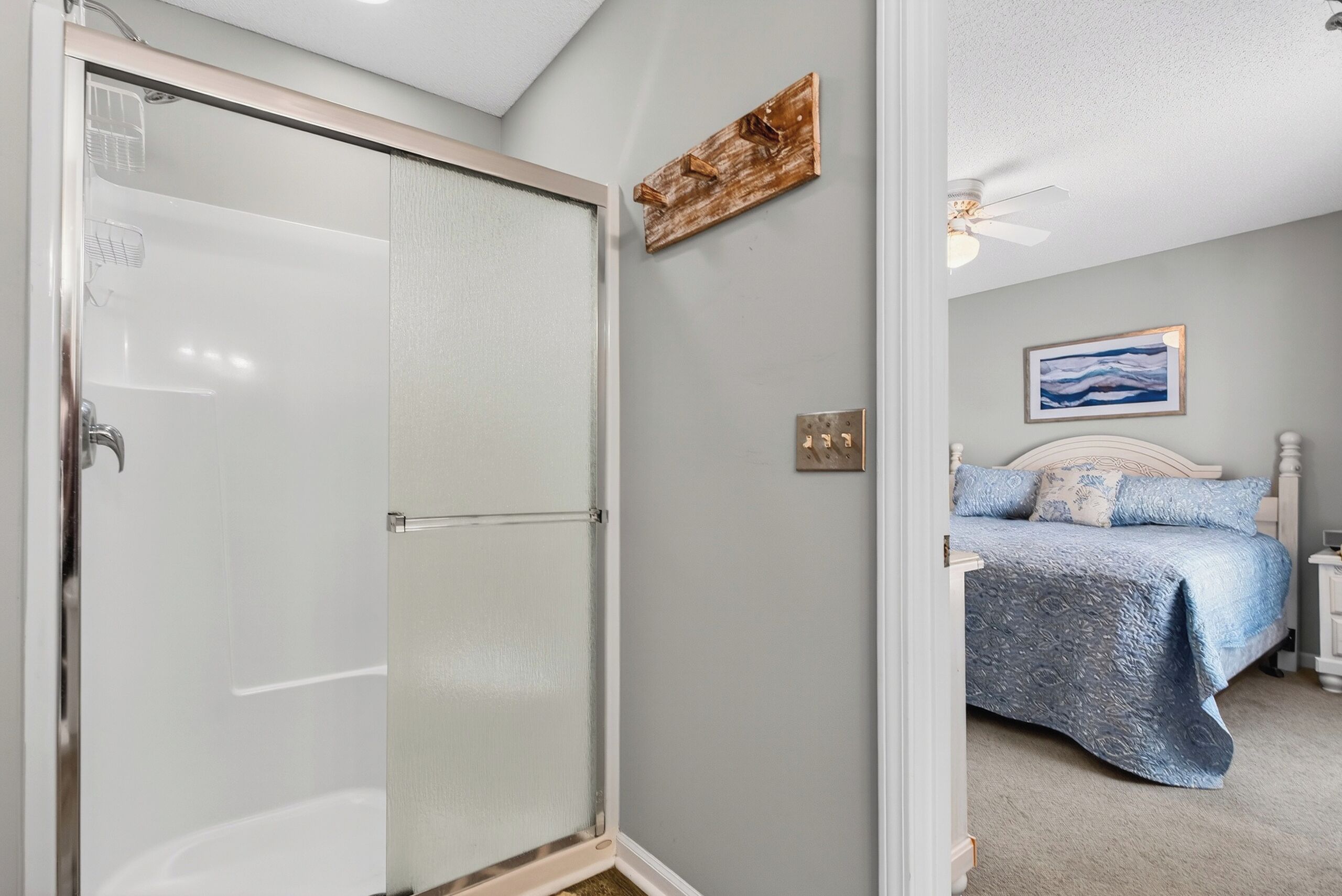 A bathroom with a glass shower door, a ceiling fan, and a bed with a blue bedspread.