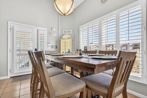A dining area with a wooden table and chairs, large windows with blinds, a chandelier, and a sign reading "BEACH BUM".