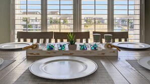 A dining table set for four with white plates, a central decorative piece, and a view through large windows. - 5BR/4BA Sleeps 10 w/ Community Pools & Short Beach Walk (Ocean Isle Beach)