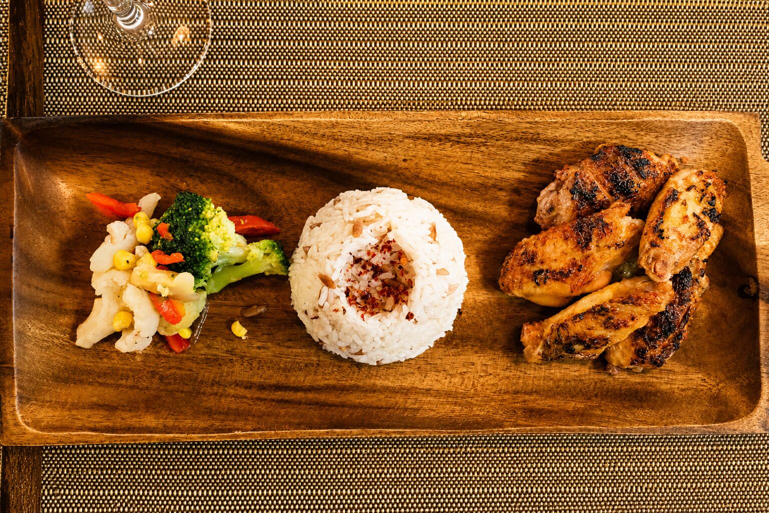 Uma bandeja de madeira com asas de frango grelhadas, uma porção de arroz e legumes variados.