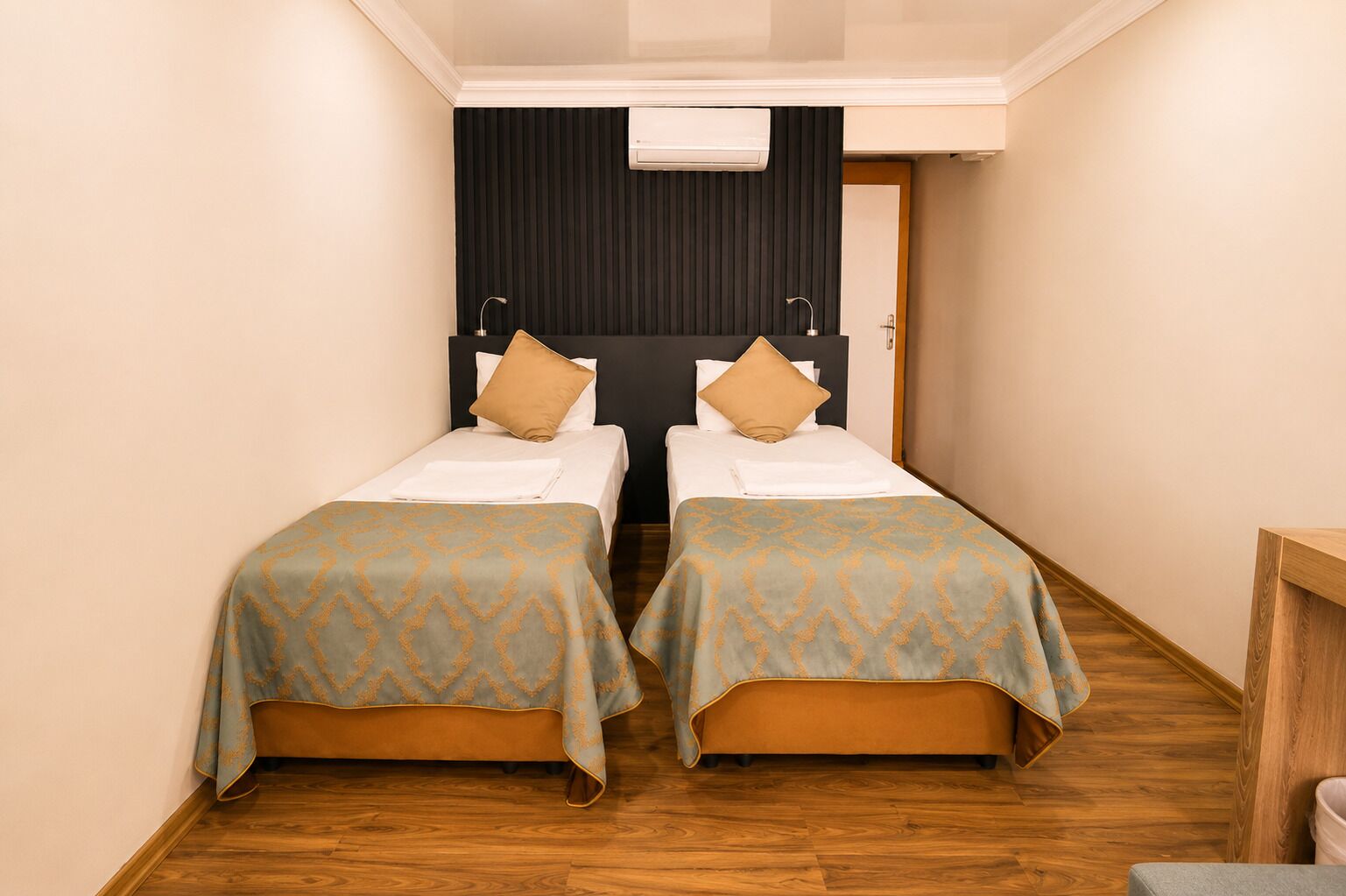 A narrow hallway leading to a room with two beds, each with a patterned bedspread and pillows, a wooden floor, and a black wall with a white air conditioning unit.