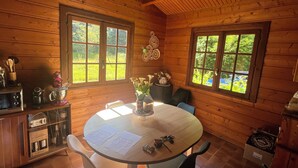 Interieur van een houten hutje met een ronde eettafel, twee stoelen, een keuken met kastjes en twee ramen met uitzicht op groen.