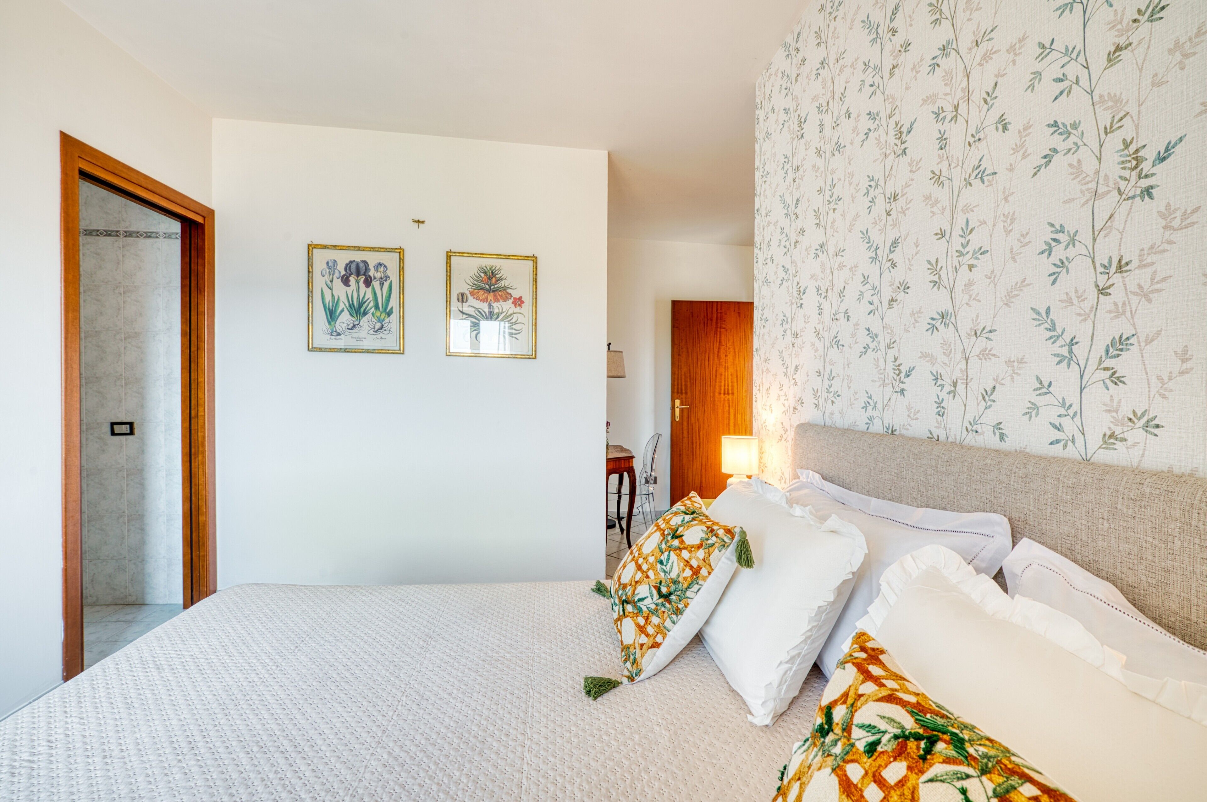 Un dormitorio con cama, almohadas, una mesita de noche, una lámpara y dos grabados botánicos enmarcados en la pared.