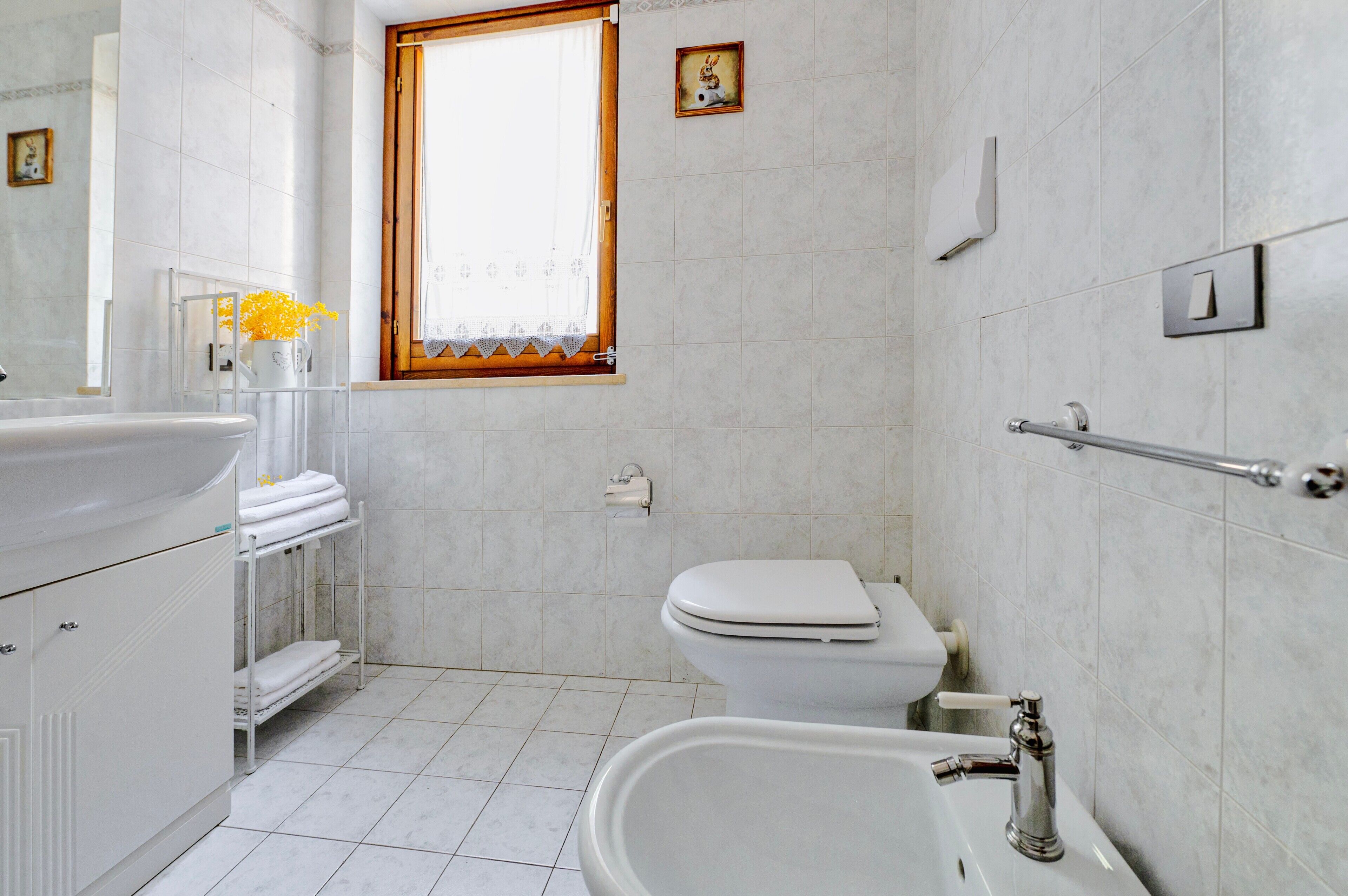 A bathroom with a toilet, sink, and bathtub. There is a window with a curtain, a towel rack, and a picture on the wall.