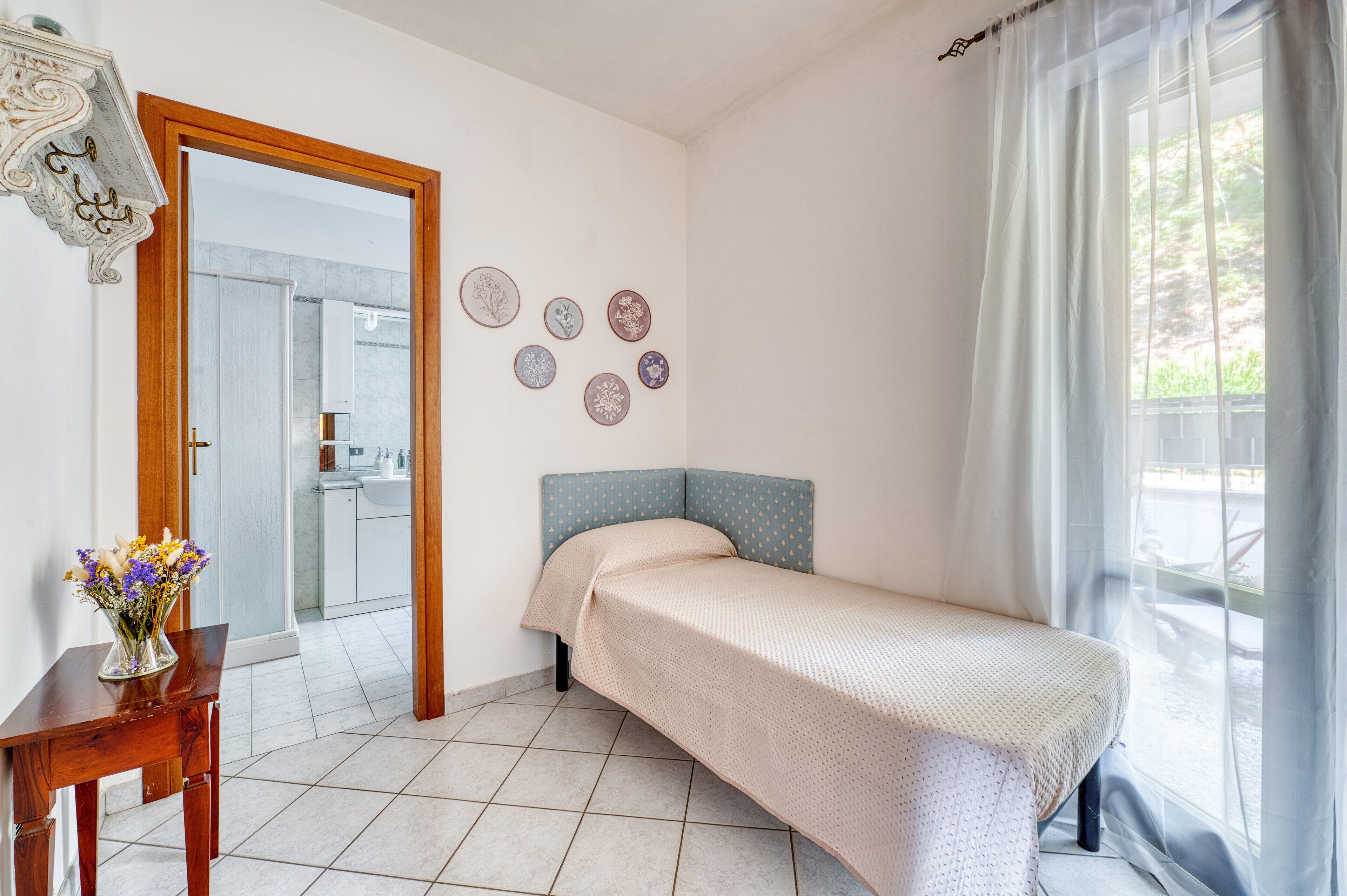 Une chambre à coucher avec un lit, une petite table sur laquelle est posé un vase de fleurs, une salle de bain visible à travers une porte ouverte, et une fenêtre ornée de rideaux légers.