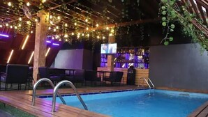 Een buitenzwembadgedeelte met een houten terras, een metalen zwembadtrap en een bar met flessen op planken.