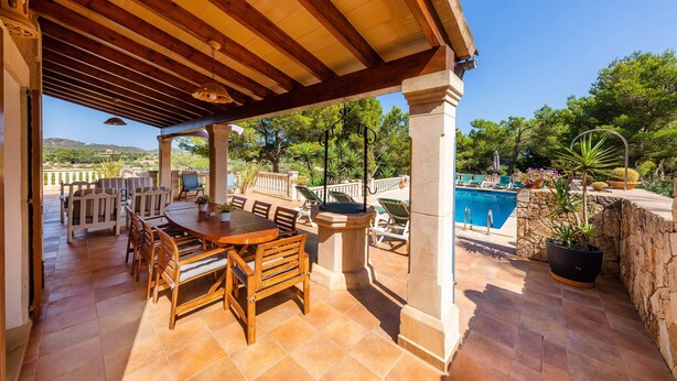Un patio al aire libre con una mesa y sillas de madera, un muro de piedra y una piscina al fondo.
