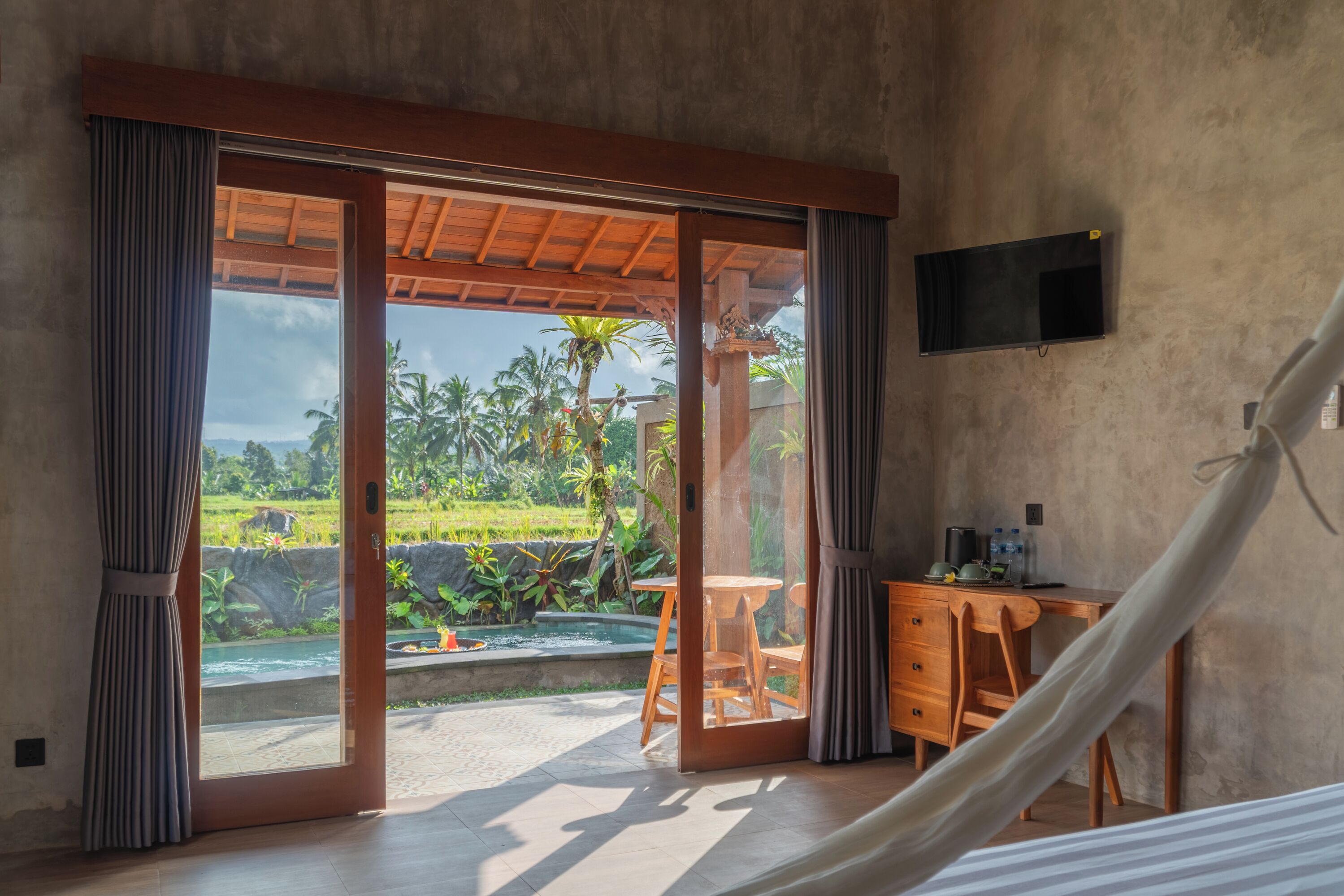 A room with a sliding glass door leading to a pool area, a wooden desk, and a TV mounted on the wall.