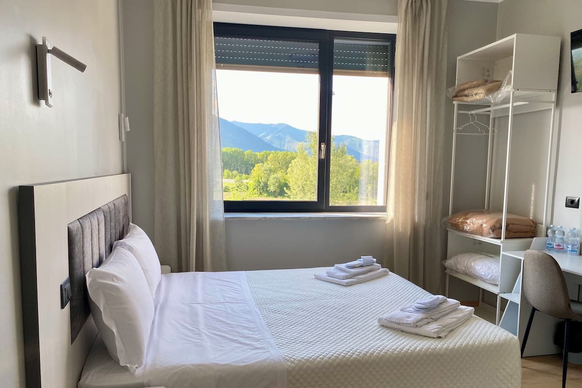 A neatly made bed with a remote control on a bedside table, a window with a view of greenery and mountains, and a white wardrobe with folded clothes and a towel.