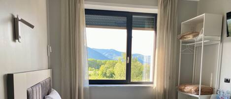 A neatly made bed with a remote control on a bedside table, a window with a view of greenery and mountains, and a white wardrobe with folded clothes and a towel.
