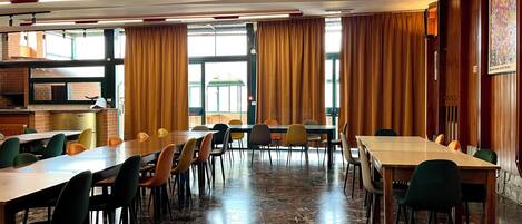 A spacious dining area with multiple tables and chairs, large windows with curtains, and a kitchen visible in the background.