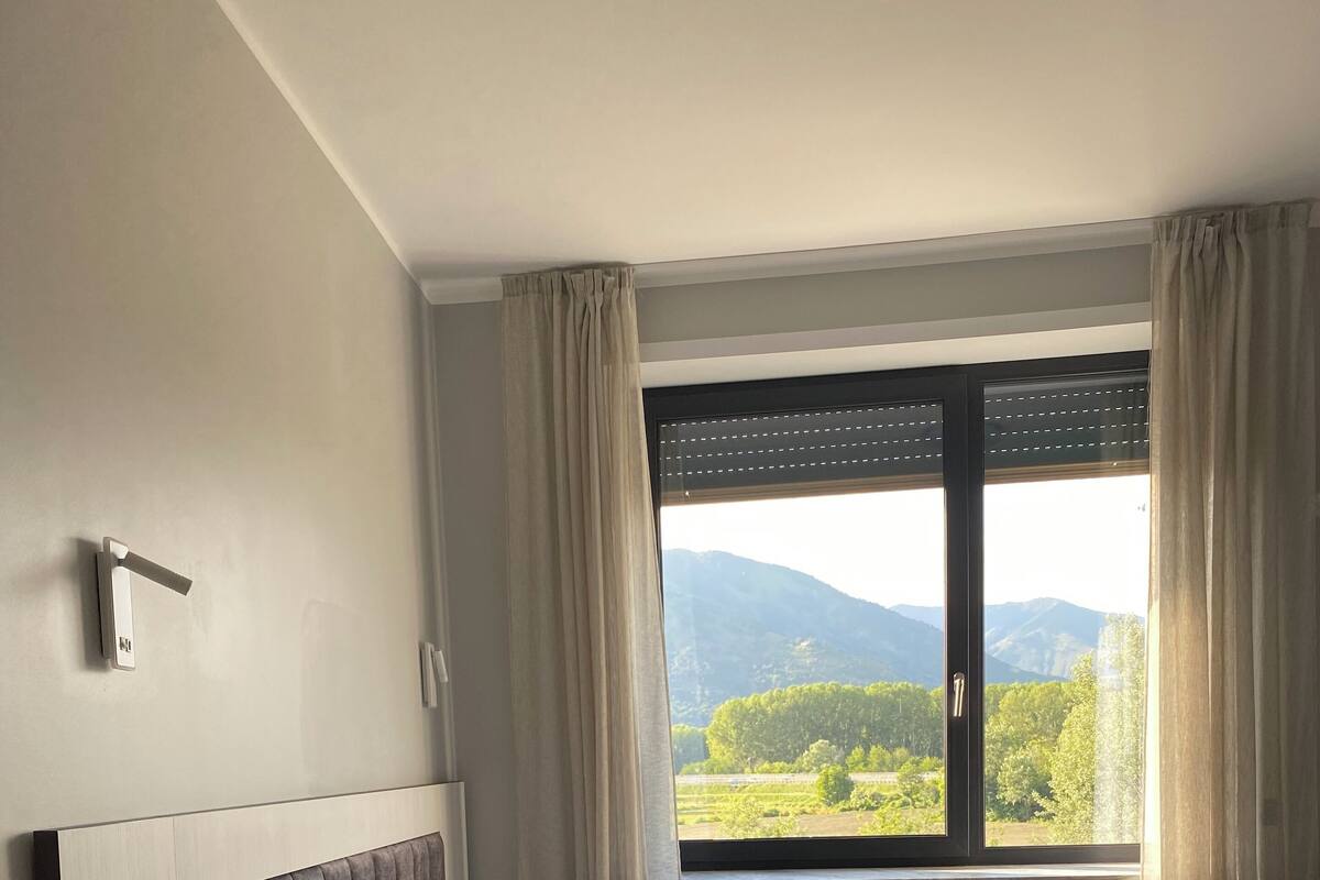 A neatly made bed with white linens and towels, a bedside table with a remote control, and a window with curtains.