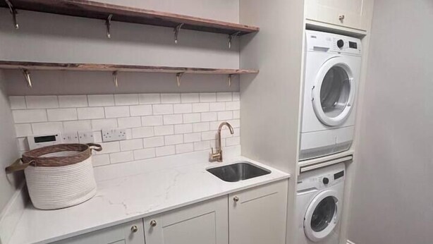 A compact kitchen with white cabinets, a built-in sink, wooden shelves, and a stacked washer and dryer. - City Victorian Townhouse Cork, Kennedy Park (Cork)