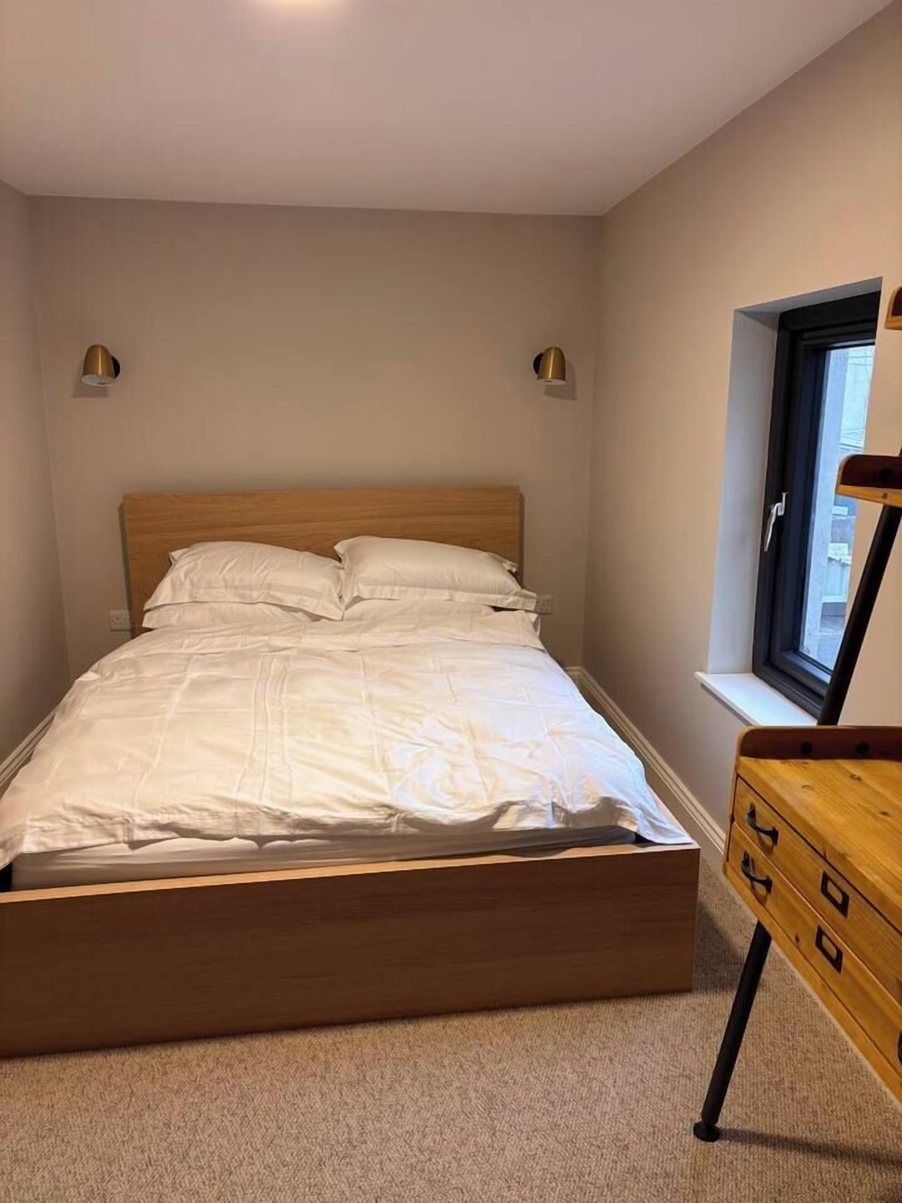 A bedroom with a wooden bed frame, white bedding, two wall-mounted lamps, and a window with a view of buildings.