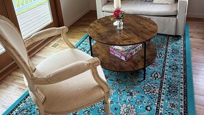 Una habitación acogedora con una silla de madera, una mesa redonda de centro con un jarrón y un libro, un sofá blanco con cojines decorativos y un cuadro en la pared.
