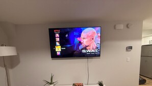 Ein Wohnzimmer mit an der Wand montiertem Flachbildfernseher, einer weißen Unterhaltungseinheit darunter und einer Stehlampe links.