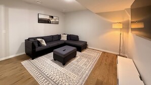 Ein modernes Wohnzimmer mit einem Ecksofa, einem Pouf, einem gemusterten Teppich und einer Stehlampe.