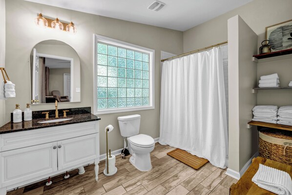 A bathroom with a white vanity, a large mirror, a window with frosted glass, a white toilet, a shower curtain, and a wooden bench.
