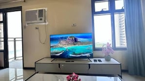 Hotellihuone, jossa on jalustalla oleva taulutelevisio, seinälle asennettu ilmastointilaite ja ikkuna, jossa on verhot.