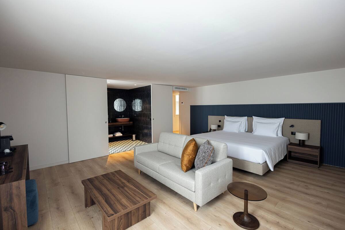 Een moderne hotelkamer met een bank, een bed, een houten bijzettafeltje en een klein nachtkastje.