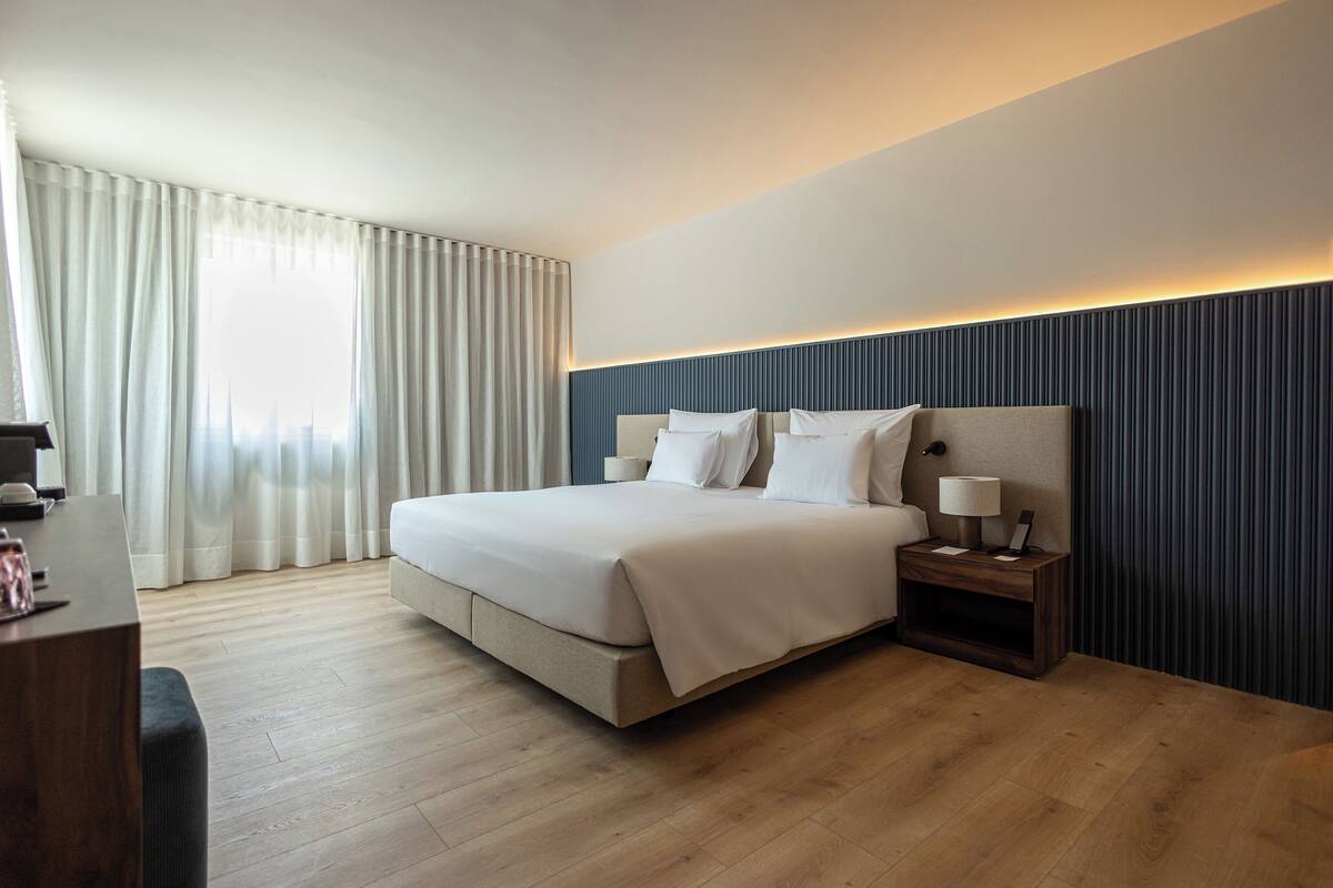 Een moderne hotelkamer met een groot bed, een nachtkastje met een lamp, en uitzicht op de stad door dunne gordijnen.