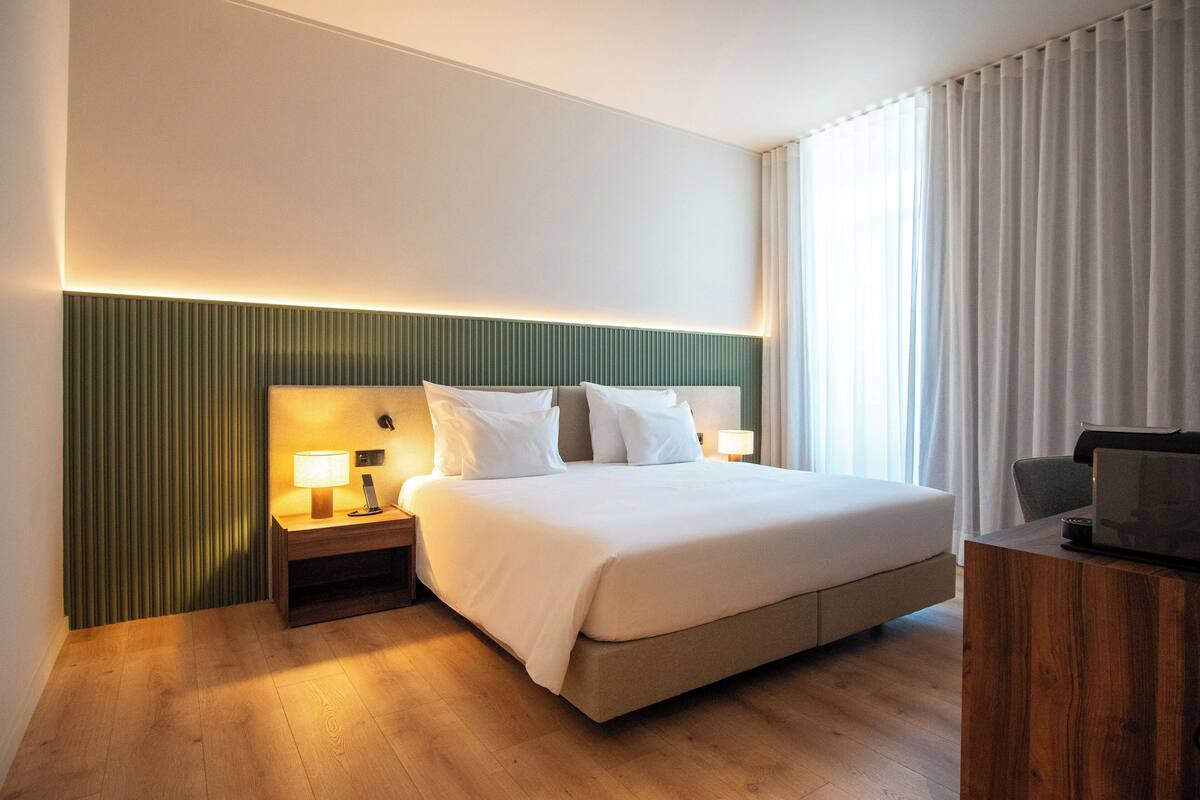 Hotelkamer met een groot bed, nachtkastjes met lampen, een houten bureau en uitzicht op het omliggende landschap door dunne gordijnen.