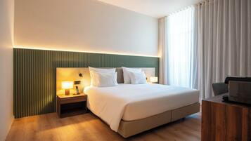A hotel room with a large bed, bedside tables with lamps, a wooden desk, and a view of the outdoors through sheer curtains.