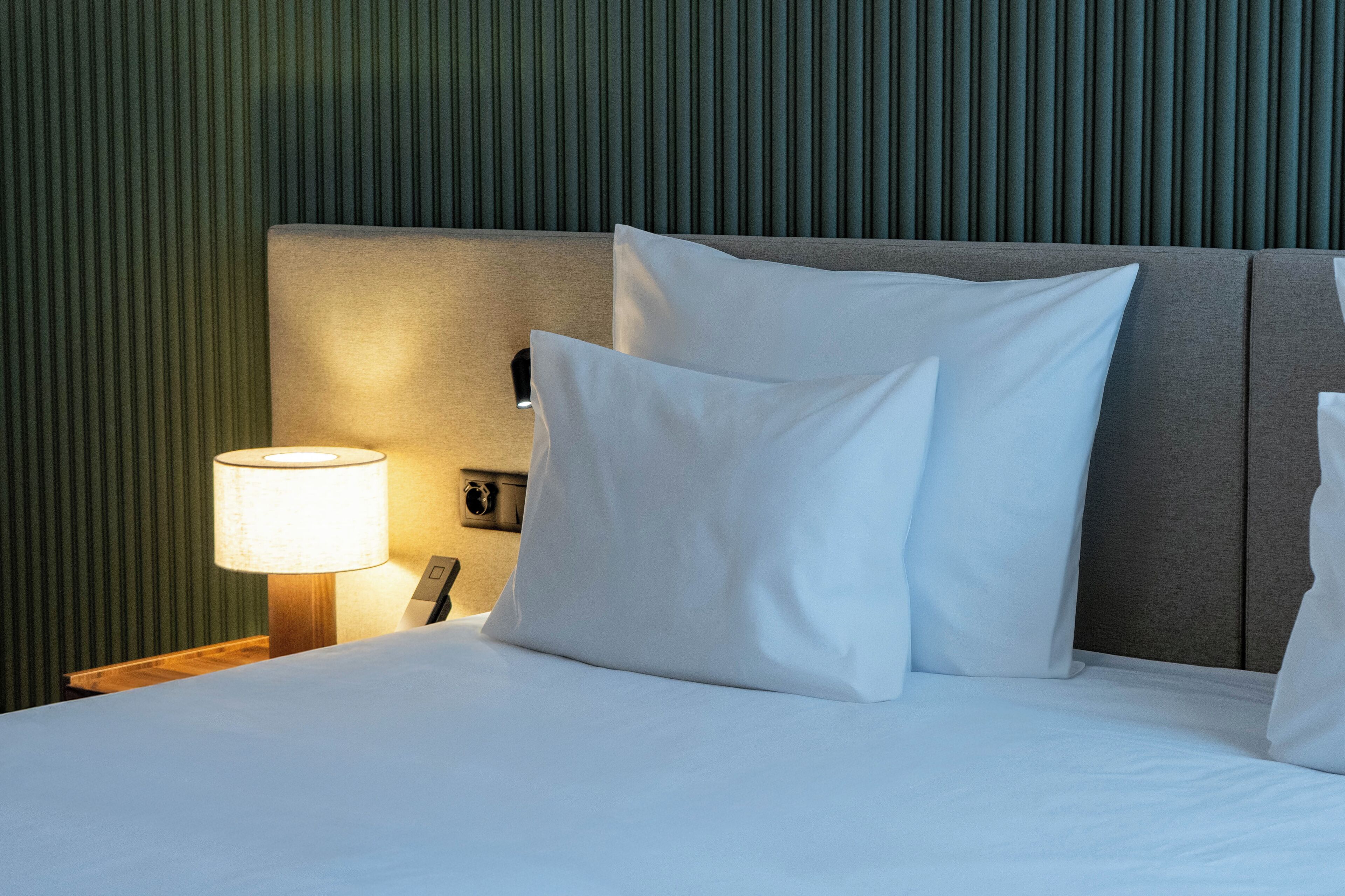 A neatly made bed with white linens and pillows, a bedside lamp, and a wall with vertical paneling.