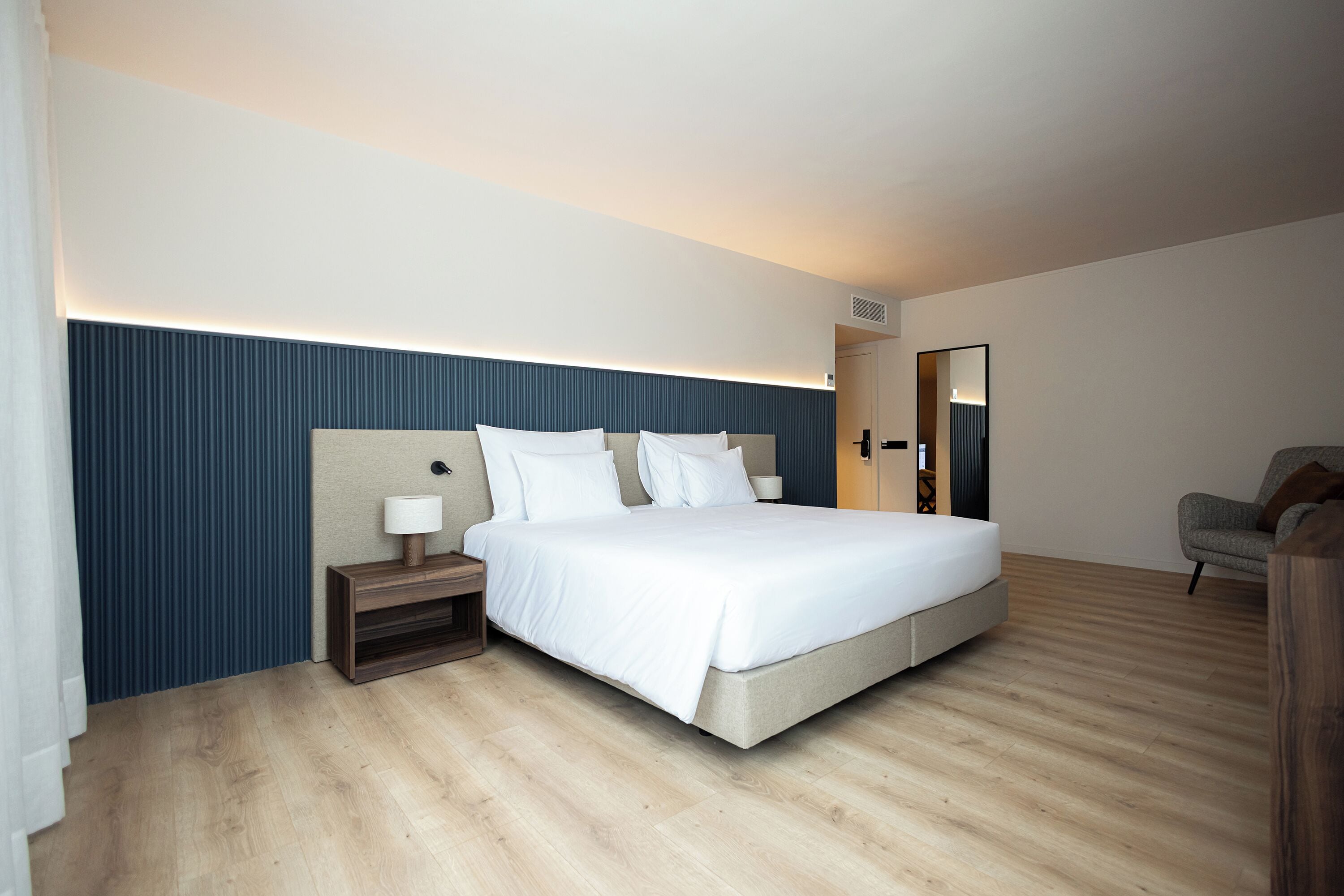 Ein modernes Hotelzimmer mit einem großen Bett, einem Nachttisch mit Lampe, Holzboden und einer dunkelblauen Akzentwand.
