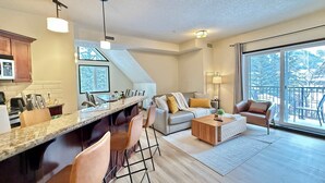 A modern kitchen with a central island, a dining area with a high table and chairs, a living room with a sofa and coffee table, and a large sliding glass door to a balcony. - Grande Rockies - Newly Renovated MTN View Retreat (Canmore)