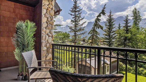 A balcony with wicker furniture, a stone wall, and a view of mountains and trees. - Grande Rockies - Newly Renovated MTN View Retreat (Canmore)