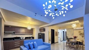 Un salon moderne avec un canapé bleu, un coin repas comprenant une table et des chaises, et une cuisine dotée de meubles sombres et d’un lustre.