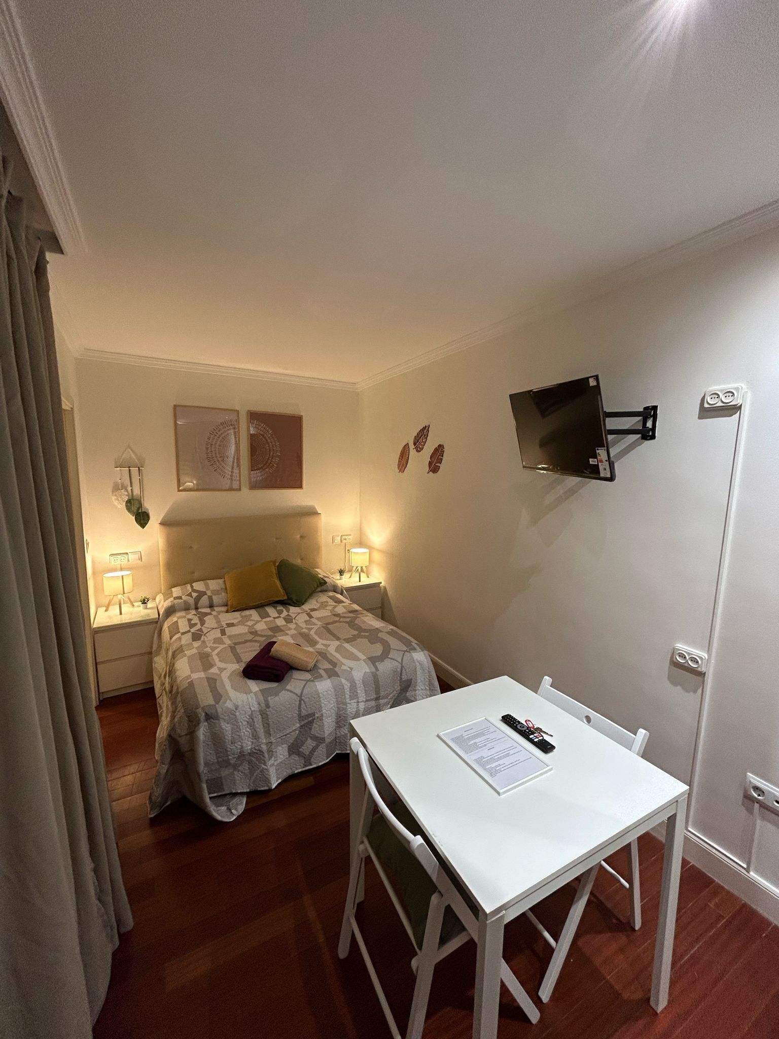 Un dormitorio pequeño y bien iluminado con cama, mesitas de noche, escritorio y televisor montado.