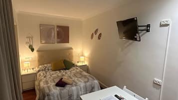Un dormitorio pequeño y bien iluminado con cama, mesitas de noche, escritorio y televisor montado.