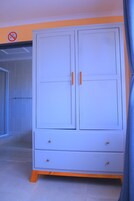 Un armario blanco con dos cajones y dos puertas, un cartel de prohibido fumar en la pared, y una cortina azul parcialmente visible.