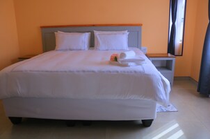 Una cama bien hecha con sábanas y almohadas blancas, una mesita de noche con lámpara y un espejo en la pared.