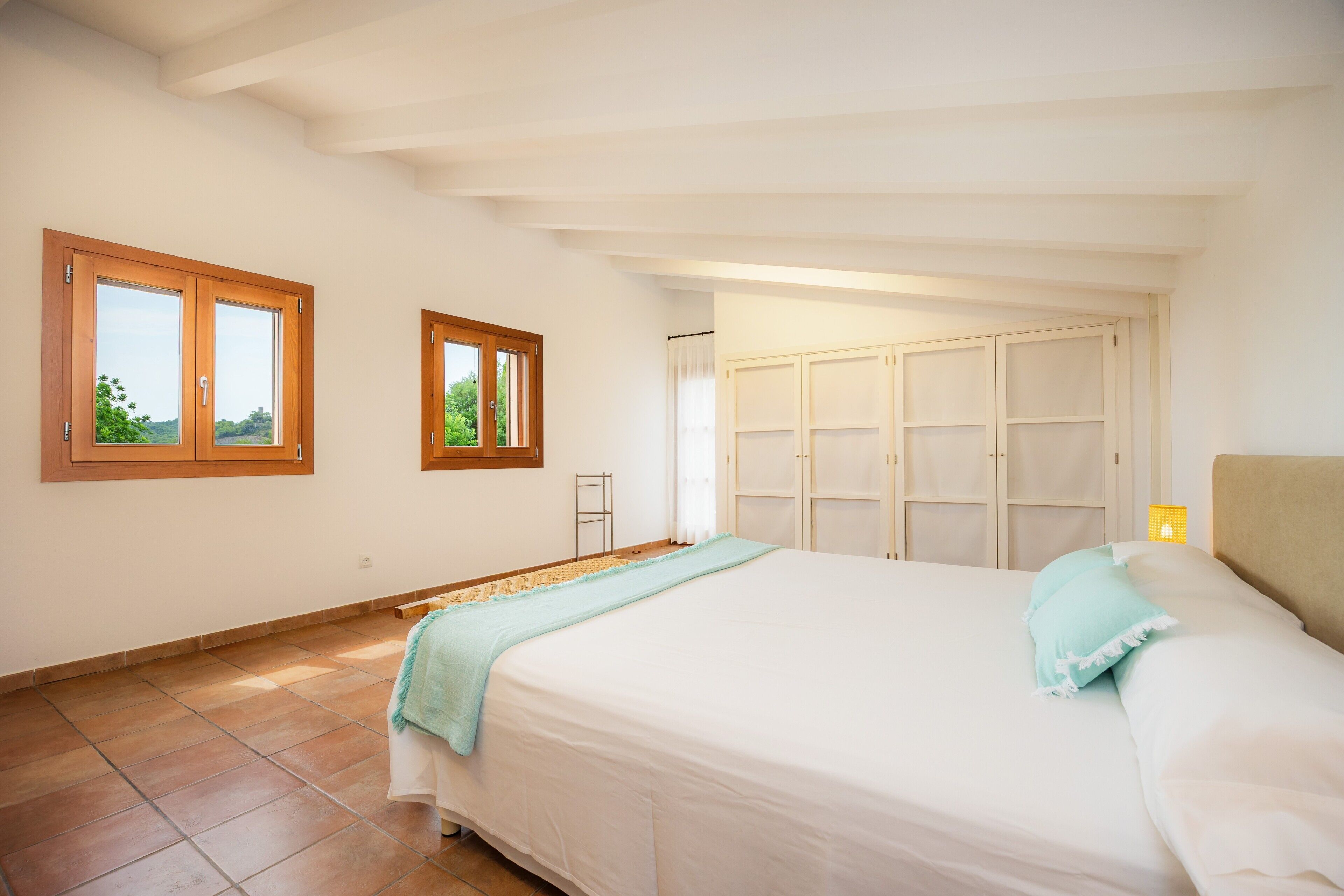 Un dormitorio con cama, dos ventanas, un armario y vistas al verdor.