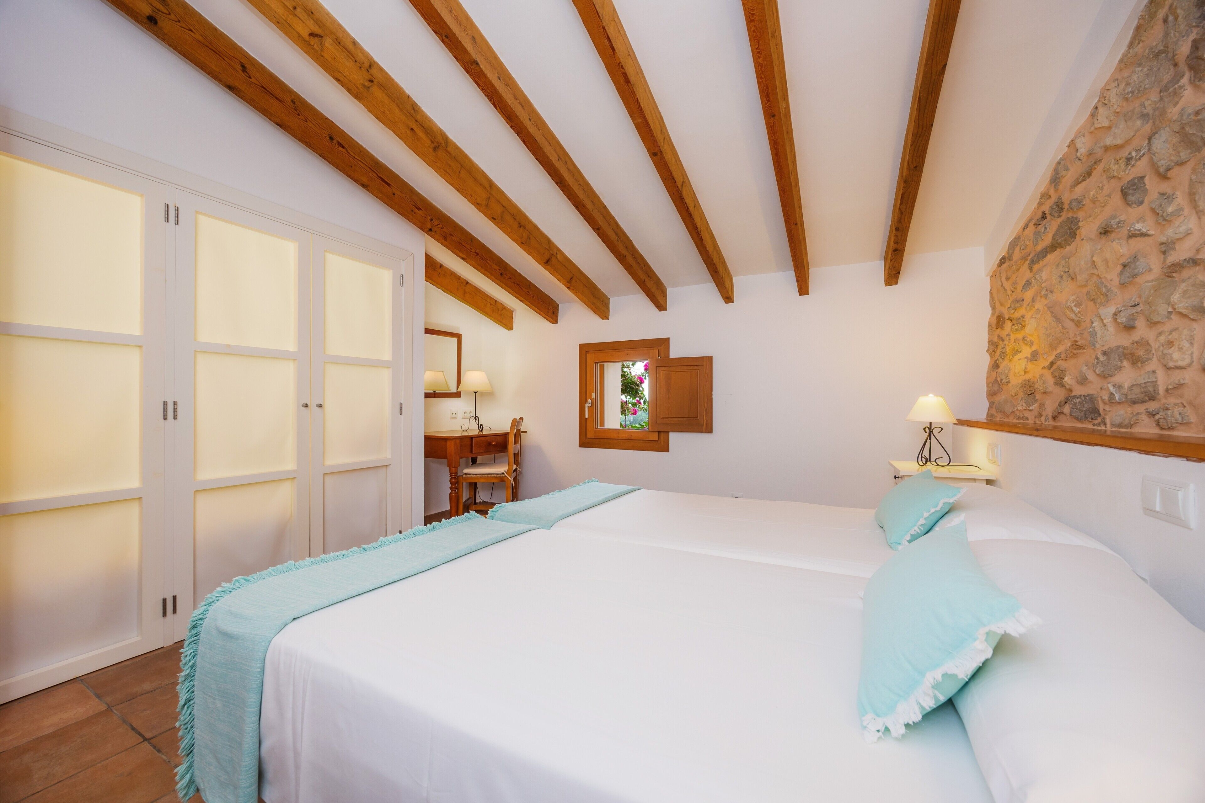 A bedroom with a bed, wooden beams, a stone wall, a desk with a chair, and a window.