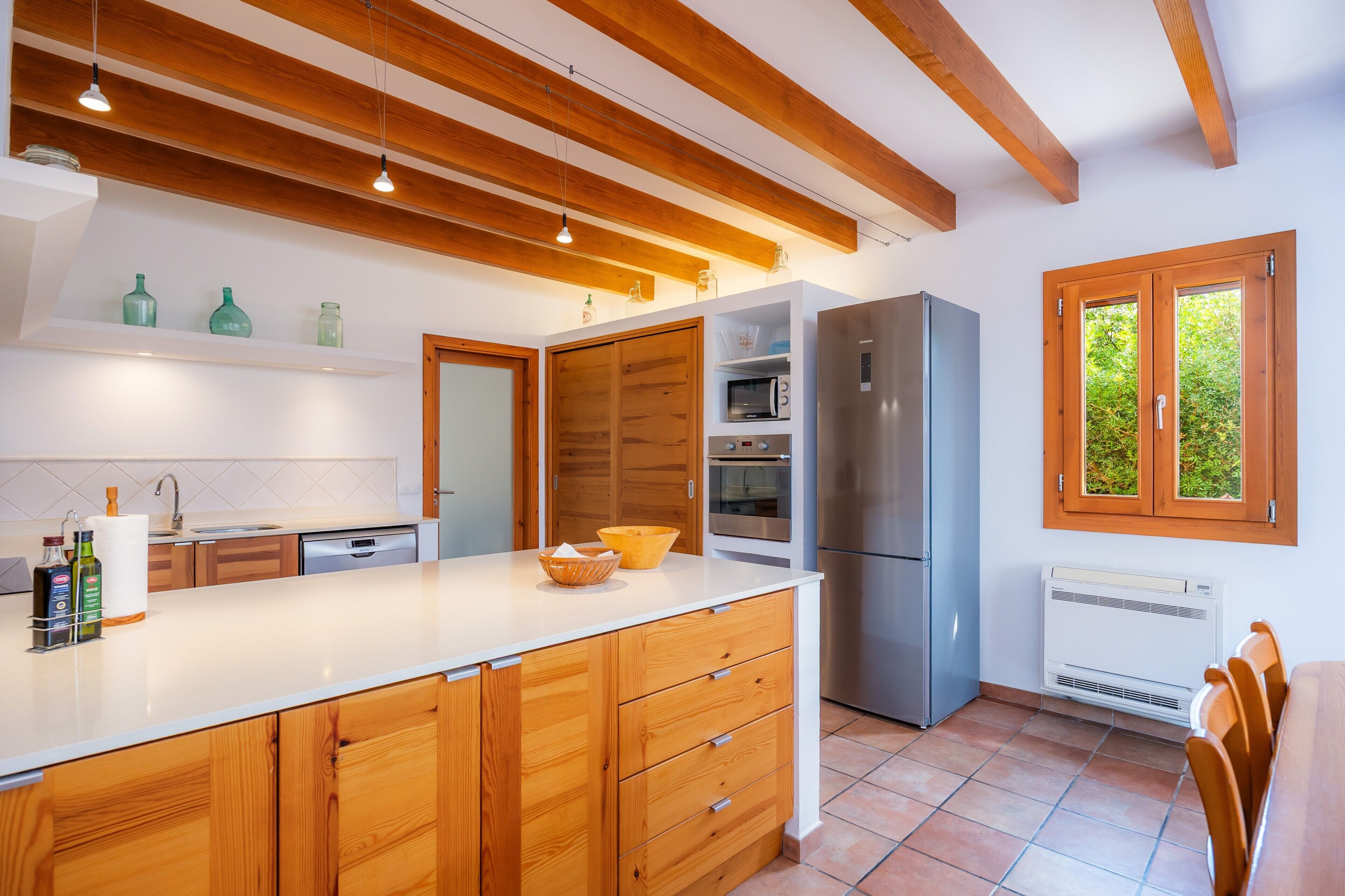 A modern kitchen with wooden cabinets, a large island, built-in appliances, and a window with a view of greenery.