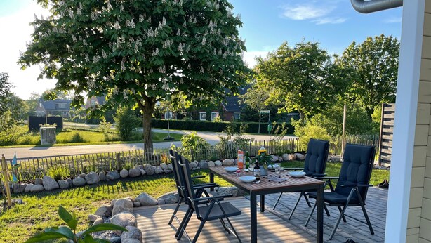 Eine Holzterrasse mit Tisch und Stühlen, umgeben von einer Steinbordüre und viel Grün.